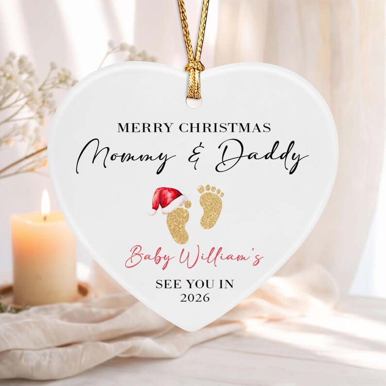 A white heart-shaped ceramic ornament hanging from a gold cord. It features the text Merry Christmas Mommy & Daddy, Baby William's, and See You In 2026, with two gold glitter baby footprints, one wearing a small red Santa hat.