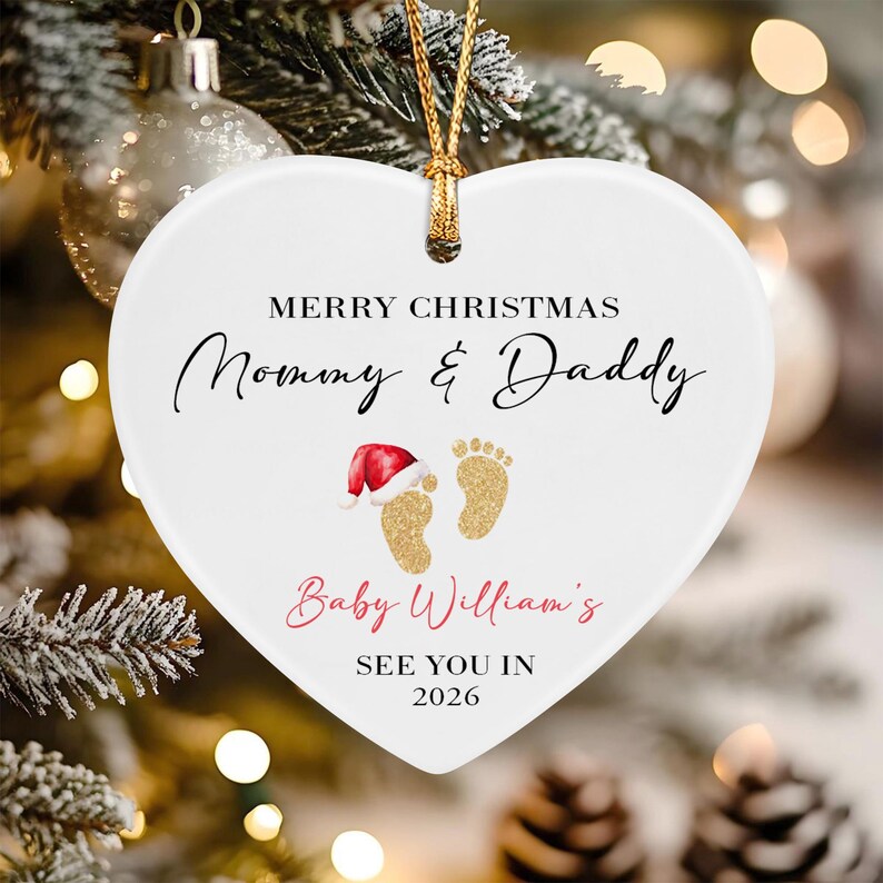 A white heart-shaped ceramic ornament with a gold string. It features the text MERRY CHRISTMAS Mommy & Daddy, Baby William's, and SEE YOU IN 2026, along with a graphic of two gold baby footprints, one wearing a red Santa hat.