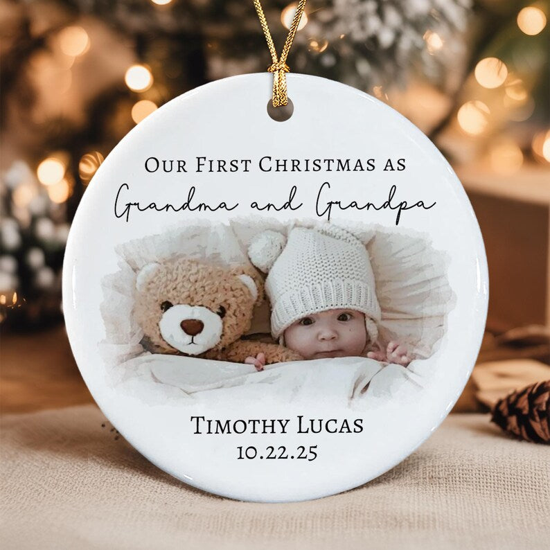 A round white ceramic ornament featuring a gold hanging string. The text reads Our First Christmas as Grandma and Grandpa, Timothy Lucas, and 10.22.25.