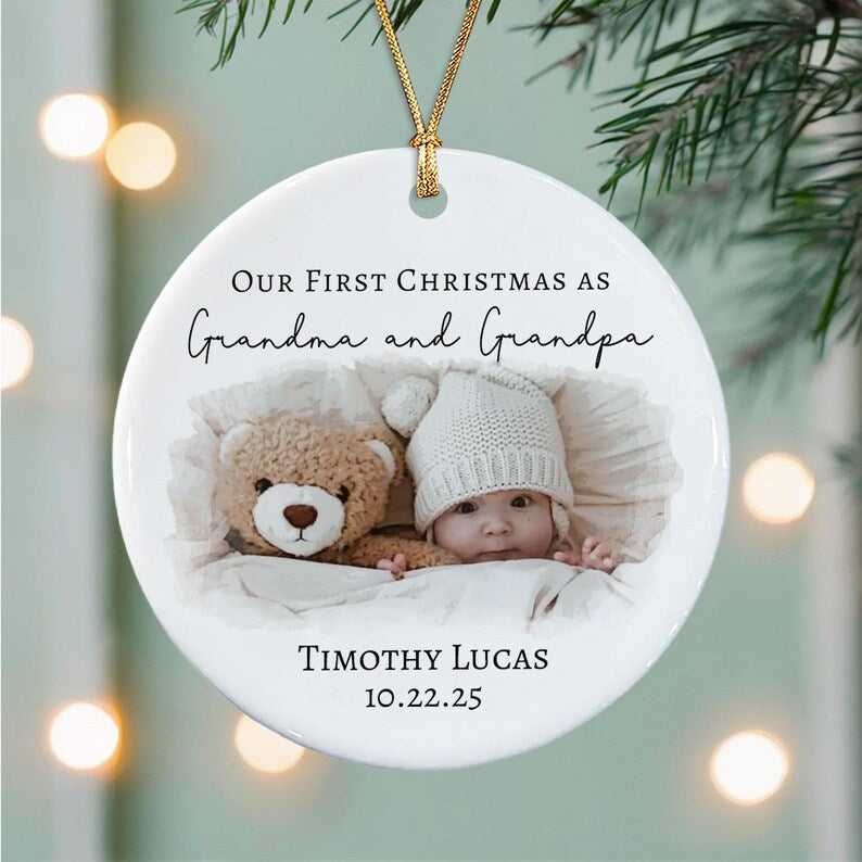A round white ceramic Christmas ornament featuring a photo of a baby in a knit hat next to a teddy bear. The text reads Our First Christmas as Grandma and Grandpa, Timothy Lucas, and the date 10.22.25. It hangs from a gold cord.