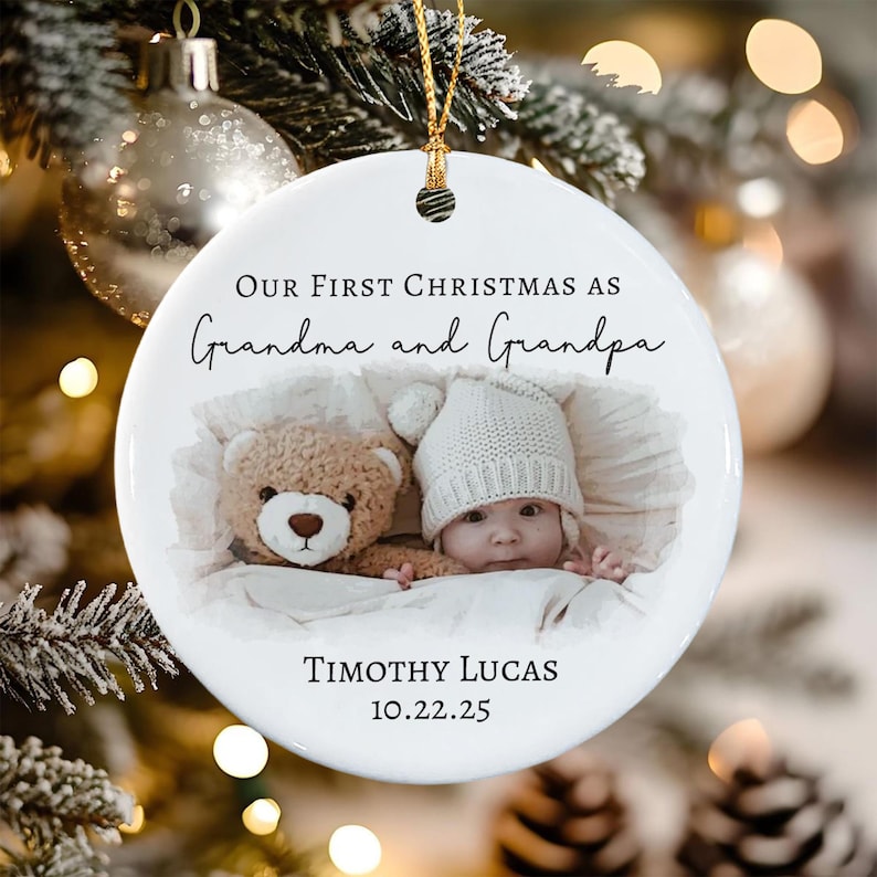 A round white ceramic ornament featuring the text Our First Christmas as Grandma and Grandpa, a photo of a baby in a white knit hat next to a brown teddy bear, and the name Timothy Lucas with the date 10.22.25 printed in black.