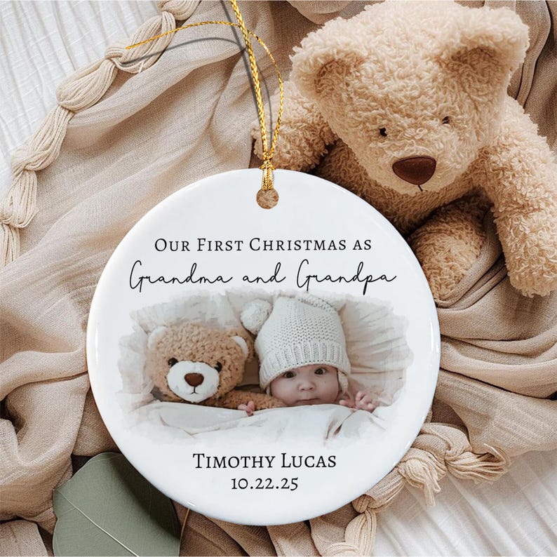 A round white ceramic ornament with a gold hanging string. The text reads "OUR FIRST CHRISTMAS AS Grandma and Grandpa" above a photo of a baby in a white knit hat next to a teddy bear.