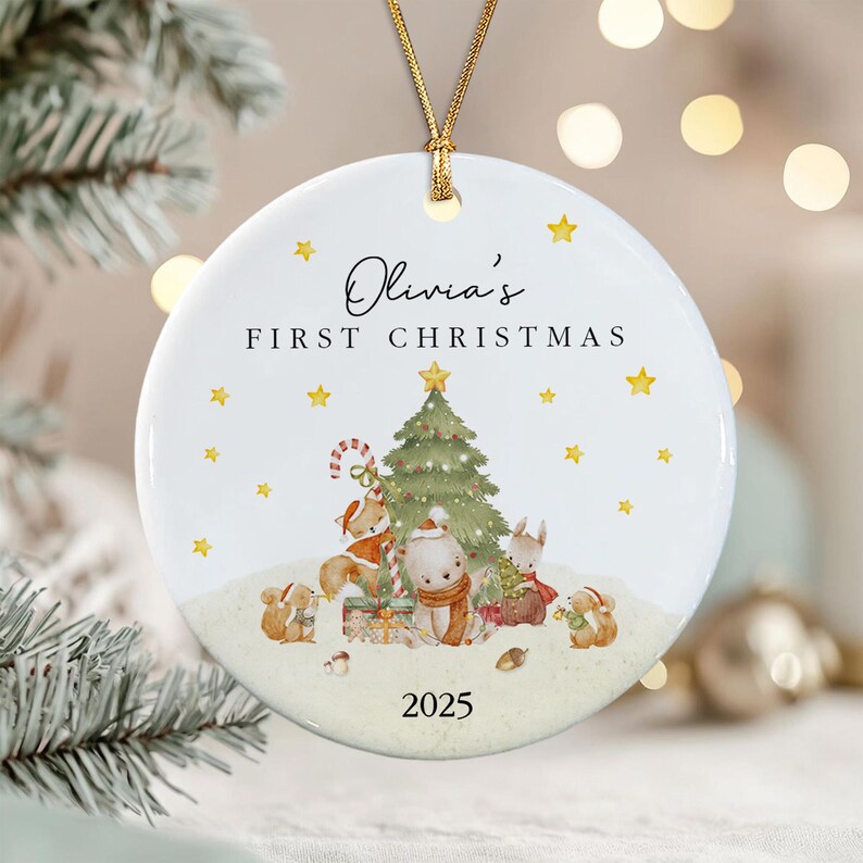 A round white ceramic Christmas ornament featuring the text Olivia's FIRST CHRISTMAS and 2025. The design includes a green Christmas tree, yellow stars, and illustrations of a fox, bear, and rabbits in winter attire.