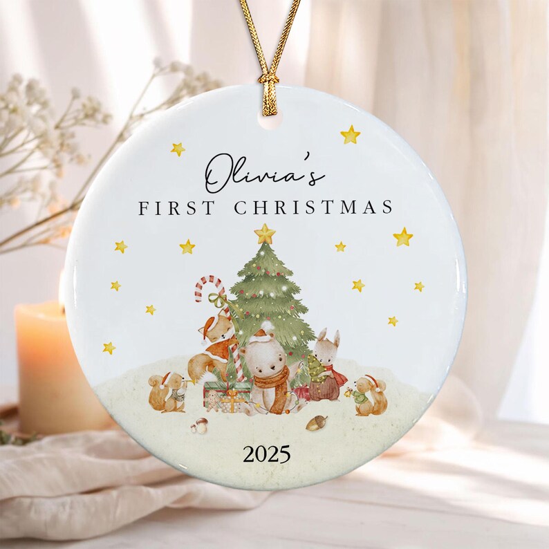 A round white ceramic ornament hanging from a gold cord. It features the text Olivia's FIRST CHRISTMAS and 2025. The design includes a green Christmas tree, yellow stars, and illustrations of a fox, bear, and squirrel.
