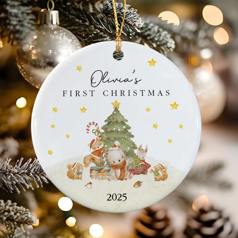 A round white ceramic Christmas ornament featuring the text Olivia's FIRST CHRISTMAS and 2025. The illustration shows a green Christmas tree surrounded by a fox, a bear, and squirrels wearing scarves and hats, with yellow stars.