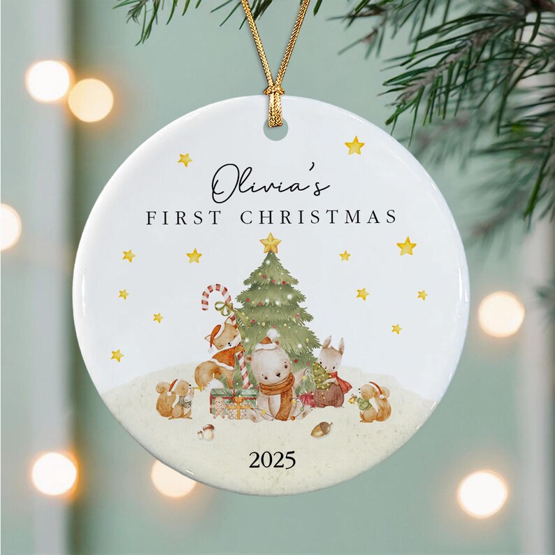 A round white ceramic Christmas ornament featuring a gold hanging cord. The design shows a green Christmas tree, woodland animals, and gifts. Text reads Olivia's FIRST CHRISTMAS and the year 2025 with yellow stars.