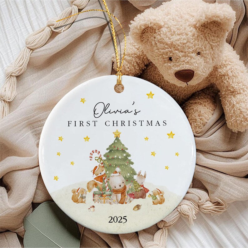 A round white ceramic ornament with a gold hanging string. The text reads Olivia's FIRST CHRISTMAS 2025. The design features a green Christmas tree, yellow stars, and illustrated woodland animals including a fox, bear, and rabbit.
