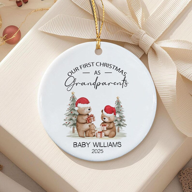 A round white ceramic Christmas ornament featuring a graphic of two adult bears and a baby bear wearing red Santa hats, holding gifts in front of pine trees. The text reads: OUR FIRST CHRISTMAS AS Grandparents BABY WILLIAMS 2025.