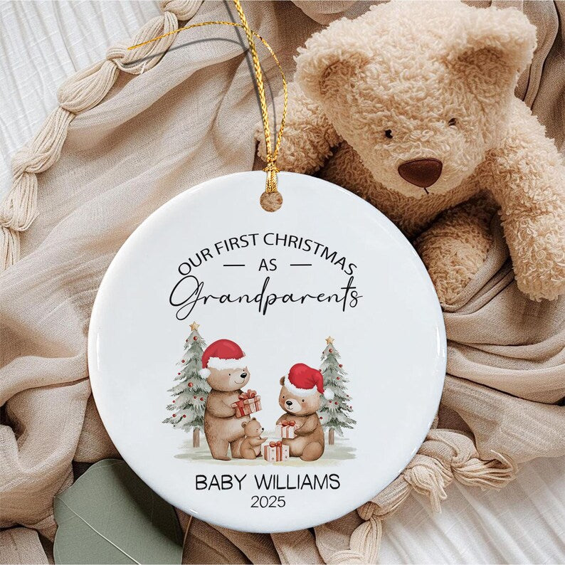 A round white ceramic ornament featuring the text "OUR FIRST CHRISTMAS AS Grandparents" and "BABY WILLIAMS 2025". The design includes two illustrated bears wearing red Santa hats, holding gifts, standing between two pine trees.