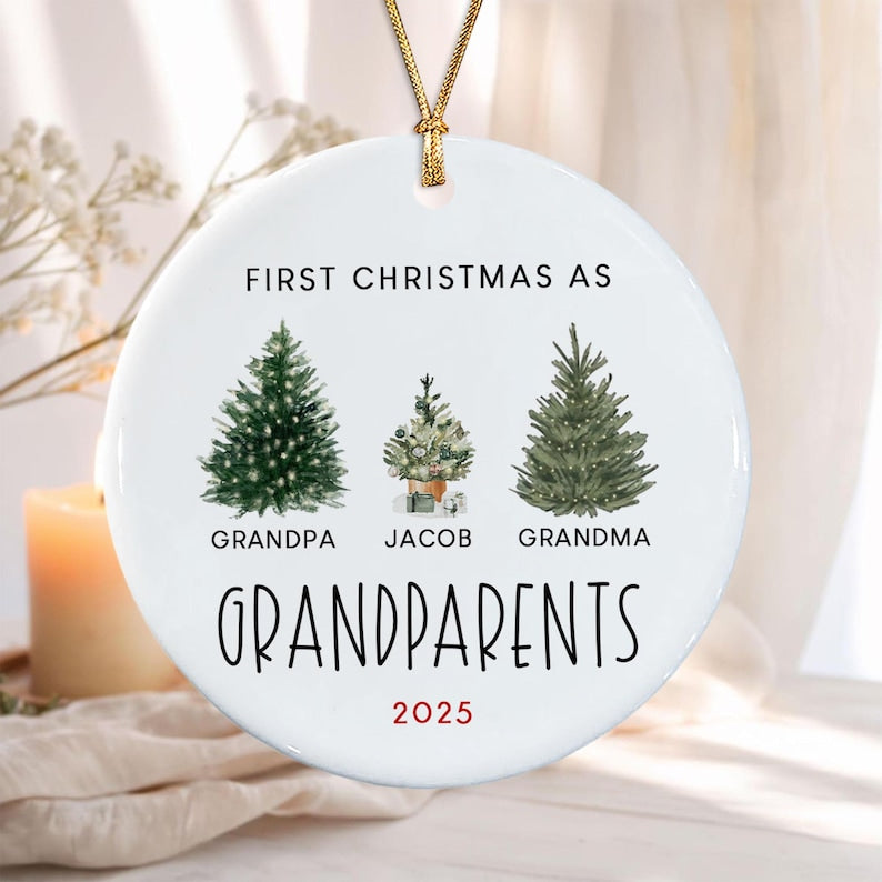 A round white ceramic ornament hanging from a gold cord. It features three illustrated evergreen trees and the text: FIRST CHRISTMAS AS GRANDPA, JACOB, GRANDMA, GRANDPARENTS, and 2025.