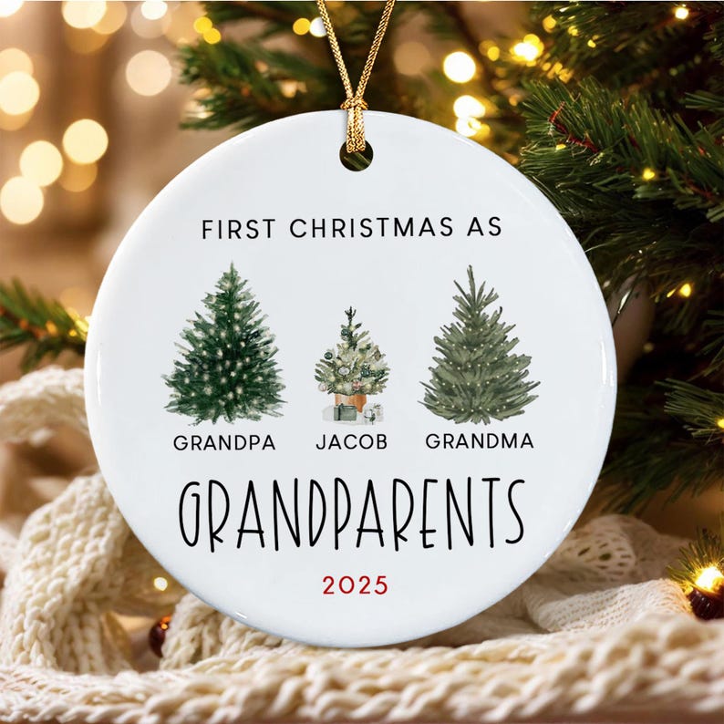A round white ceramic ornament with a gold hanging cord. The text reads FIRST CHRISTMAS AS GRANDPARENTS 2025. It features illustrations of two large pine trees and one smaller tree labeled GRANDPA, JACOB, and GRANDMA.