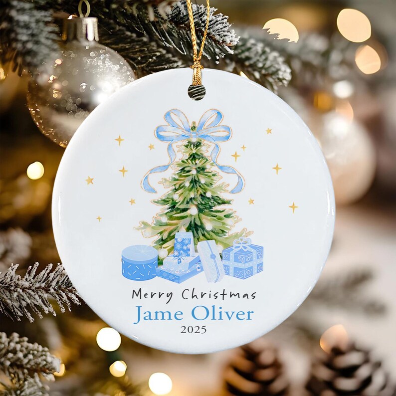A round white ceramic Christmas ornament featuring a watercolor illustration of a green pine tree topped with a light blue bow, surrounded by blue gift boxes and gold stars. The text reads Merry Christmas, Jame Oliver, 2025.