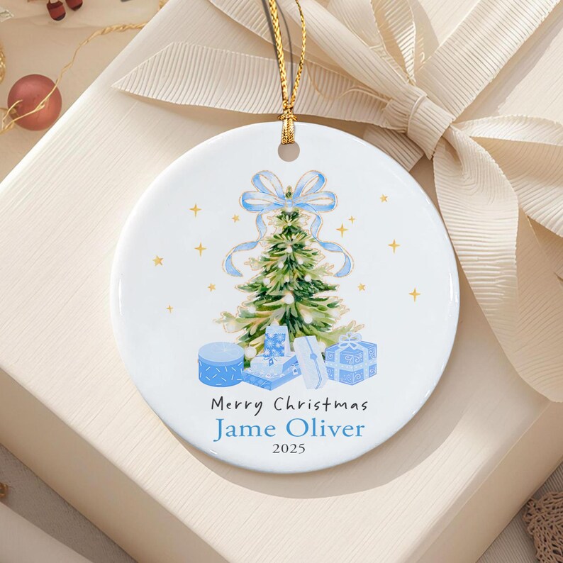 A round white ceramic ornament featuring a watercolor illustration of a green Christmas tree topped with a light blue bow, surrounded by blue gift boxes and gold stars. The text reads Merry Christmas, Jame Oliver, and 2025.