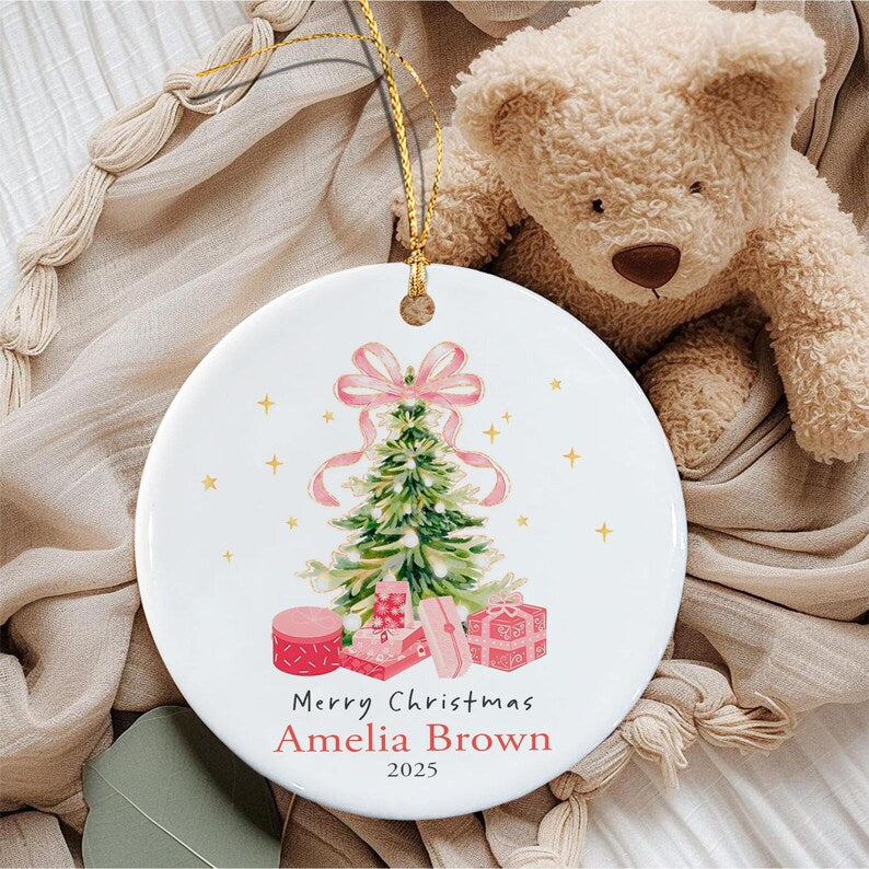 A round white ceramic ornament featuring a watercolor illustration of a green Christmas tree topped with a pink bow, surrounded by pink gift boxes. The text reads Merry Christmas, Amelia Brown, and 2025 in black and red lettering.