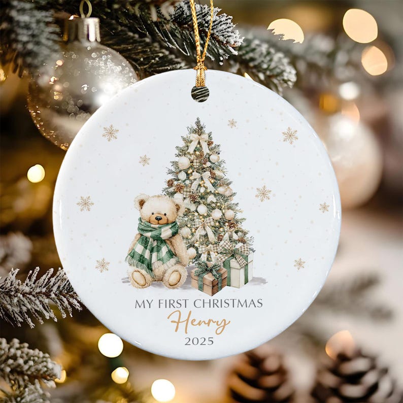 A round white ceramic ornament featuring a teddy bear wearing a green and white plaid scarf sitting next to a decorated Christmas tree and two gift boxes. The text reads MY FIRST CHRISTMAS Henry 2025 in black and gold script.