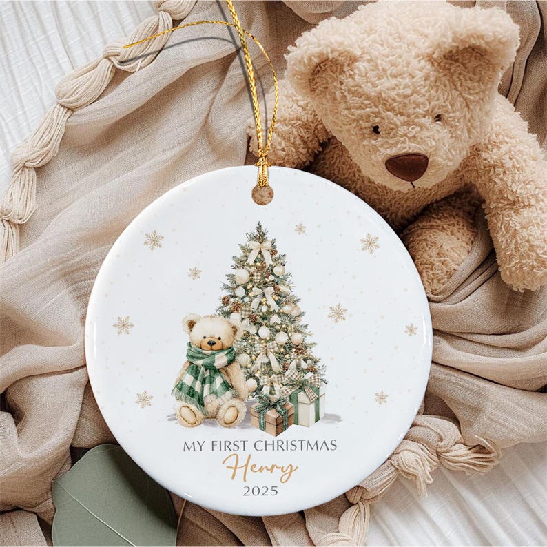 A round white ceramic ornament featuring a printed illustration of a teddy bear wearing a green and white plaid scarf sitting next to a decorated Christmas tree and gift boxes. The text reads MY FIRST CHRISTMAS Henry 2025.
