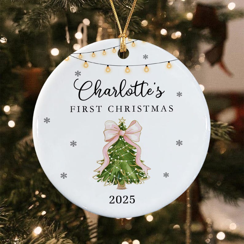 A round white ceramic Christmas ornament featuring a string of lights at the top, the text "Charlotte's FIRST CHRISTMAS", a green Christmas tree with a large pink bow, small snowflakes, and the year "2025" printed in black.