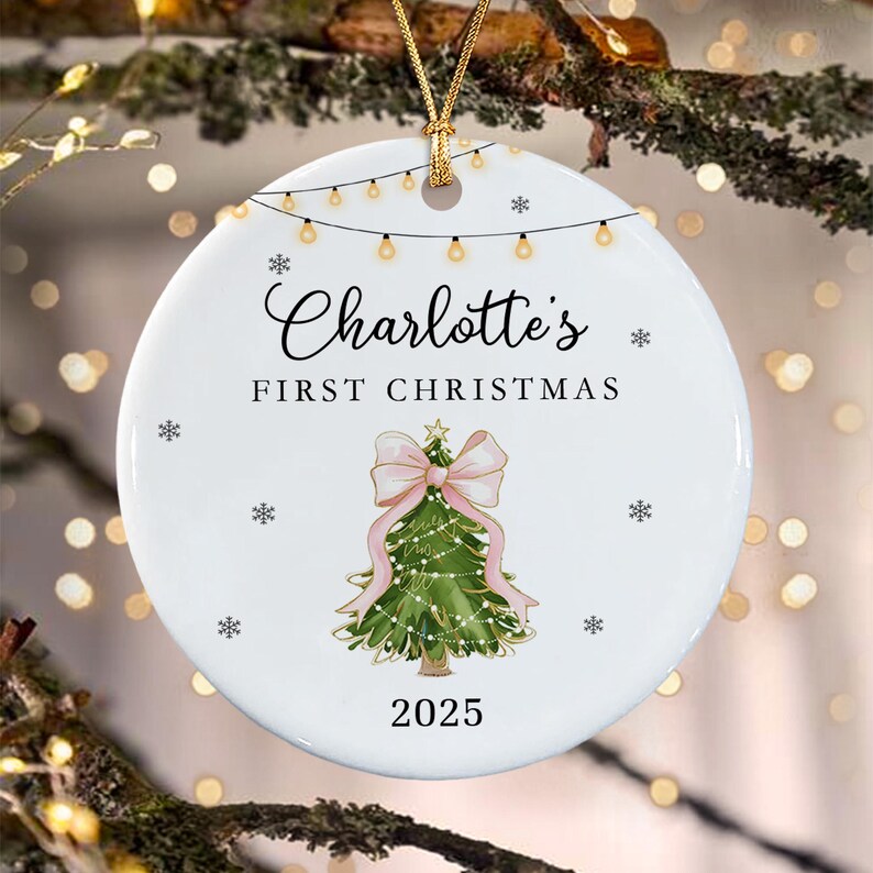 A round white ceramic ornament featuring a string of lights at the top, the text Charlotte's FIRST CHRISTMAS, a green Christmas tree with a large pink bow, and the year 2025, accented with small black snowflakes and a gold cord.