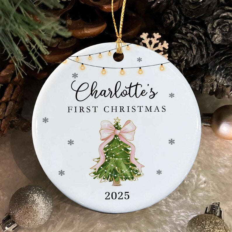 A round white ceramic Christmas ornament featuring a string of lights at the top, the text Charlotte's FIRST CHRISTMAS, a green Christmas tree with a large pink bow, and the year 2025, suspended by a gold cord for hanging.