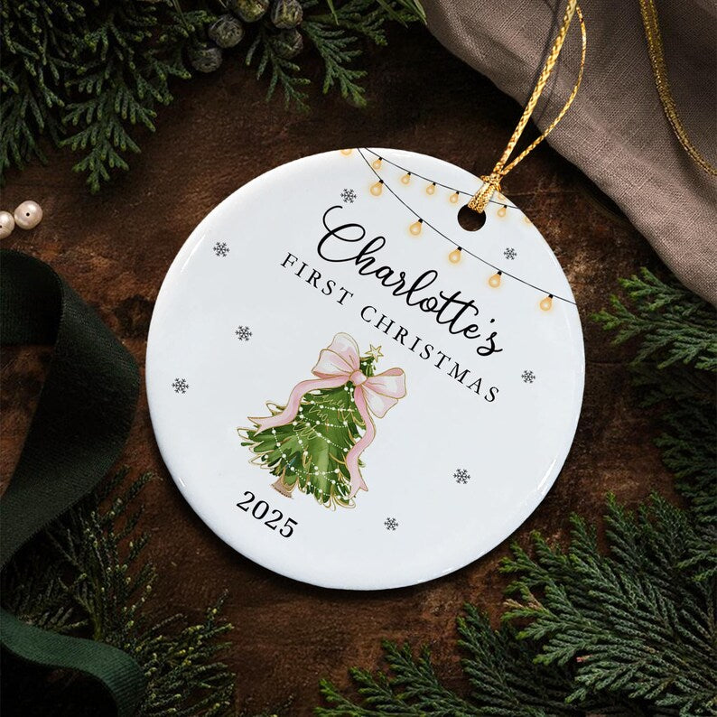 A round white ceramic ornament featuring a string of yellow fairy lights at the top, the text Charlotte's FIRST CHRISTMAS, a green Christmas tree illustration with a large pink bow, and the year 2025, suspended by a gold cord.