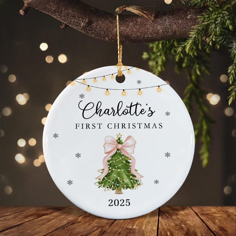A round white ceramic ornament featuring a string of lights at the top, the text "Charlotte's FIRST CHRISTMAS", a green Christmas tree illustration with a large pink bow, and the year "2025" at the bottom.