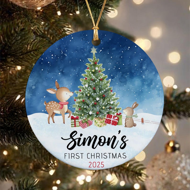 A round ceramic ornament featuring a watercolor illustration of a deer and a rabbit standing by a decorated Christmas tree in the snow. The text reads Simon's FIRST CHRISTMAS 2025. It hangs from a gold string.