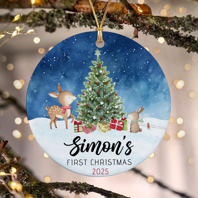 A round ceramic Christmas ornament featuring a watercolor illustration of a deer and a rabbit beside a decorated tree in the snow. The text reads Simon's FIRST CHRISTMAS 2025 in black and red lettering with a gold hanging cord.