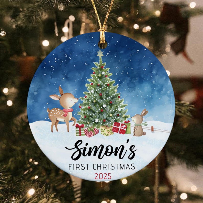 A round ceramic Christmas ornament featuring a blue snowy background with a decorated pine tree, a fawn, and a rabbit. The text reads Simon's FIRST CHRISTMAS 2025 in black and red lettering. It hangs from a gold string.