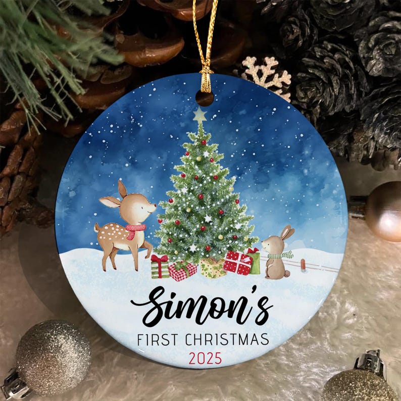 A round ceramic Christmas ornament featuring a blue snowy background with a decorated pine tree, a fawn, and a rabbit. The text reads Simon's FIRST CHRISTMAS 2025 in black and red lettering. A gold string is attached to the top.