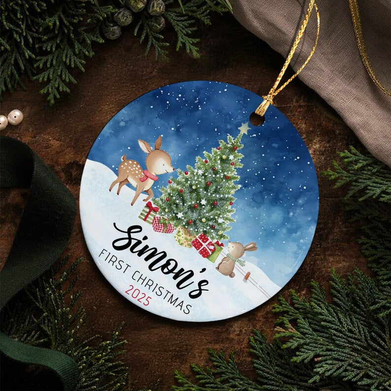A round ceramic Christmas ornament featuring a watercolor illustration of a deer and a rabbit near a decorated pine tree on a snowy hill against a dark blue starry sky. The text reads Simon's FIRST CHRISTMAS 2025.