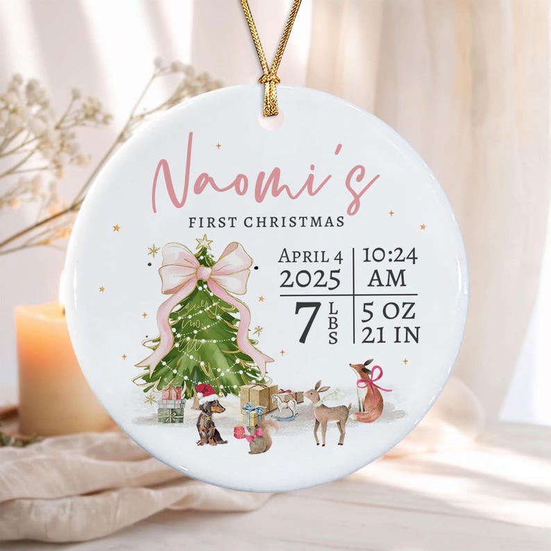 A round white ceramic ornament with a gold string featuring the text Naomi's First Christmas, a green Christmas tree with a pink bow, and illustrations of a dachshund, squirrel, deer, and fox, plus birth details: April 4 2025.