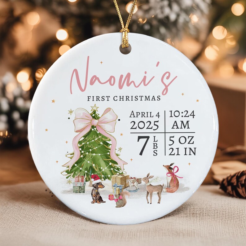A round white ceramic ornament with a gold string, featuring the text Naomi's First Christmas, birth date April 4 2025, time 10:24 AM, weight 7 lbs 5 oz, and length 21 in.