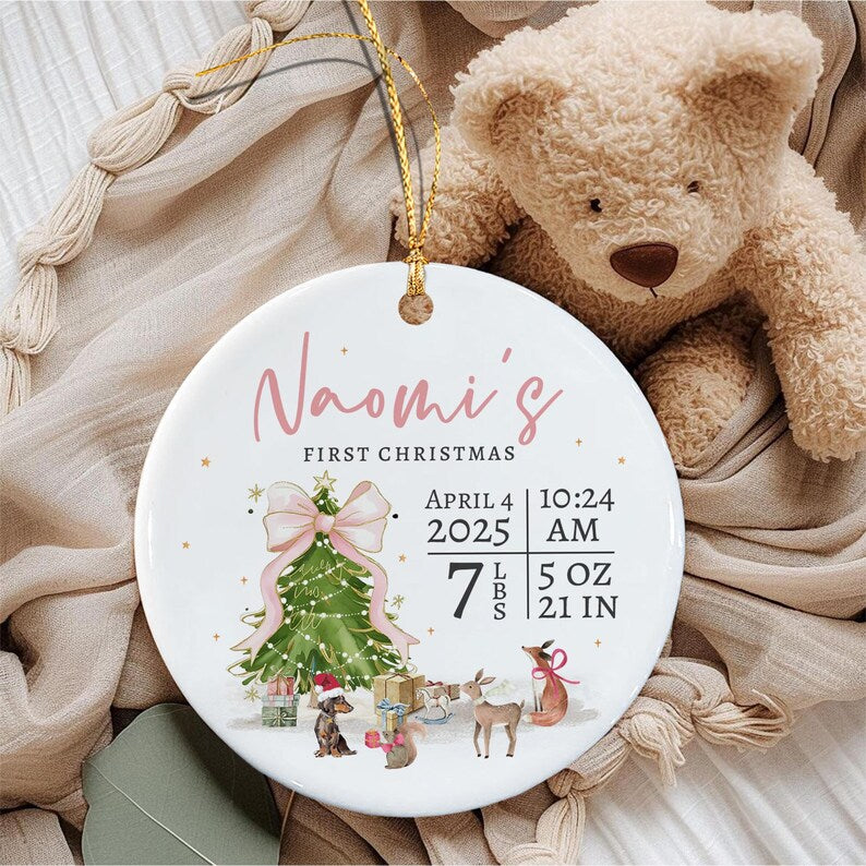 A round white ceramic ornament featuring the text Naomi's First Christmas, a green Christmas tree with a pink bow, and illustrations of a dog, a squirrel, and a deer.