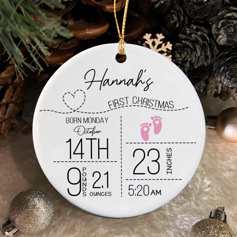 A round white ceramic ornament featuring the text Hannah's First Christmas, a dashed line with a heart, and baby birth details: Born Monday October 14th, 9 pounds 2.1 ounces, 23 inches, and 5:20 AM.