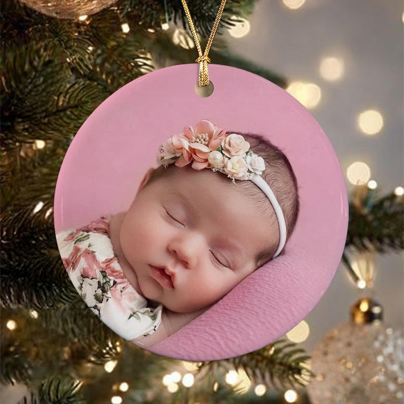 A round ceramic ornament featuring a photograph of a sleeping infant wearing a white floral headband. The baby is dressed in a white garment with a pink floral pattern. The ornament has a gold string loop for hanging on a tree.