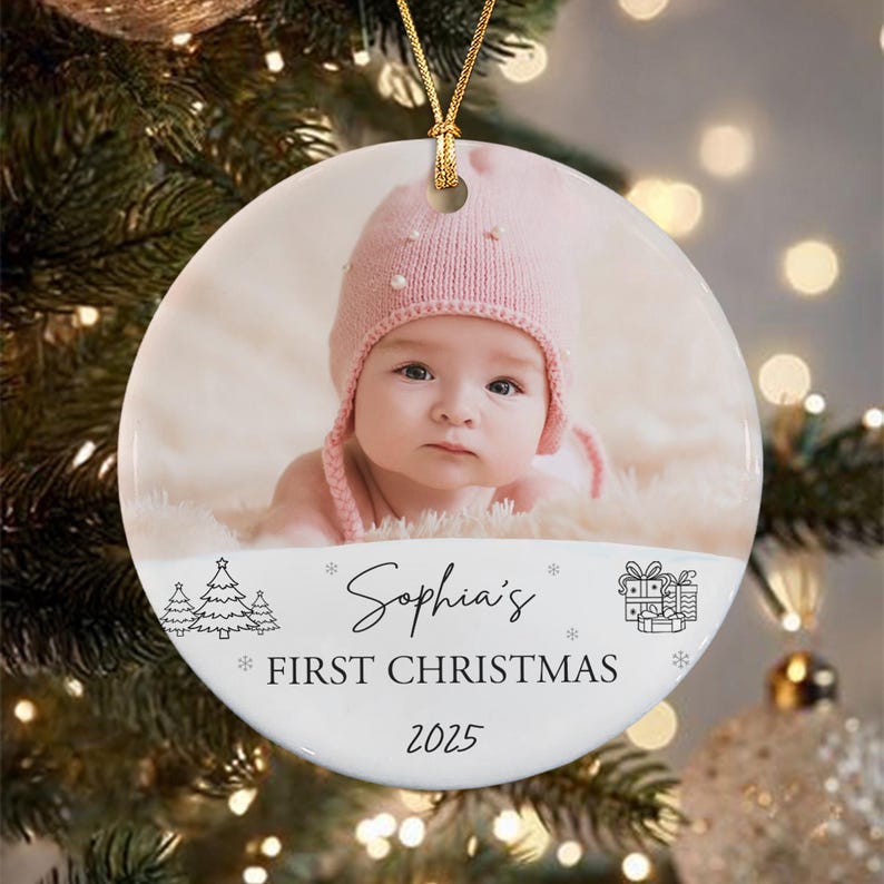 A round, white Christmas ornament with a photo of a baby wearing a pink knit hat. The ornament has the text "Sophia's First Christmas 2025" with decorative elements of trees, snowflakes, and a gift box.
