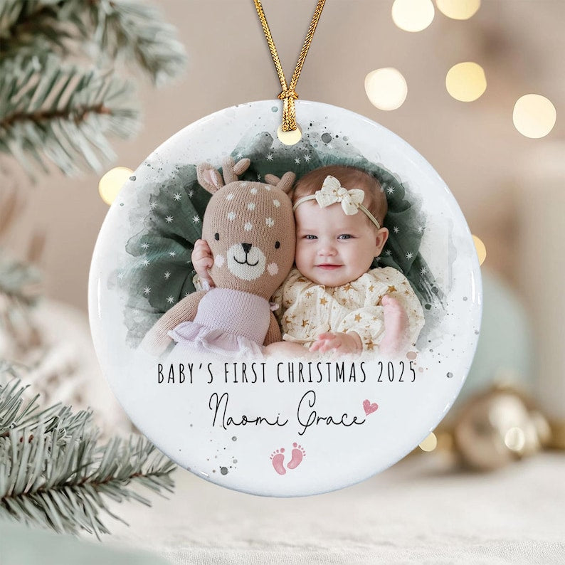 A round white ceramic ornament featuring a photo of a baby holding a plush deer toy. The text reads BABY'S FIRST CHRISTMAS 2025 and the name Naomi Grace, accompanied by a small pink heart and two tiny pink baby footprint icons.