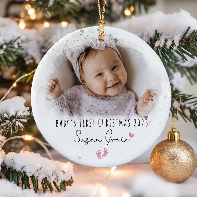 A round white ceramic ornament featuring a photograph of a baby wearing a headband and a textured sweater. The text reads BABY'S FIRST CHRISTMAS 2025 and Susan Grace, accompanied by a small pink heart and two pink baby footprints.