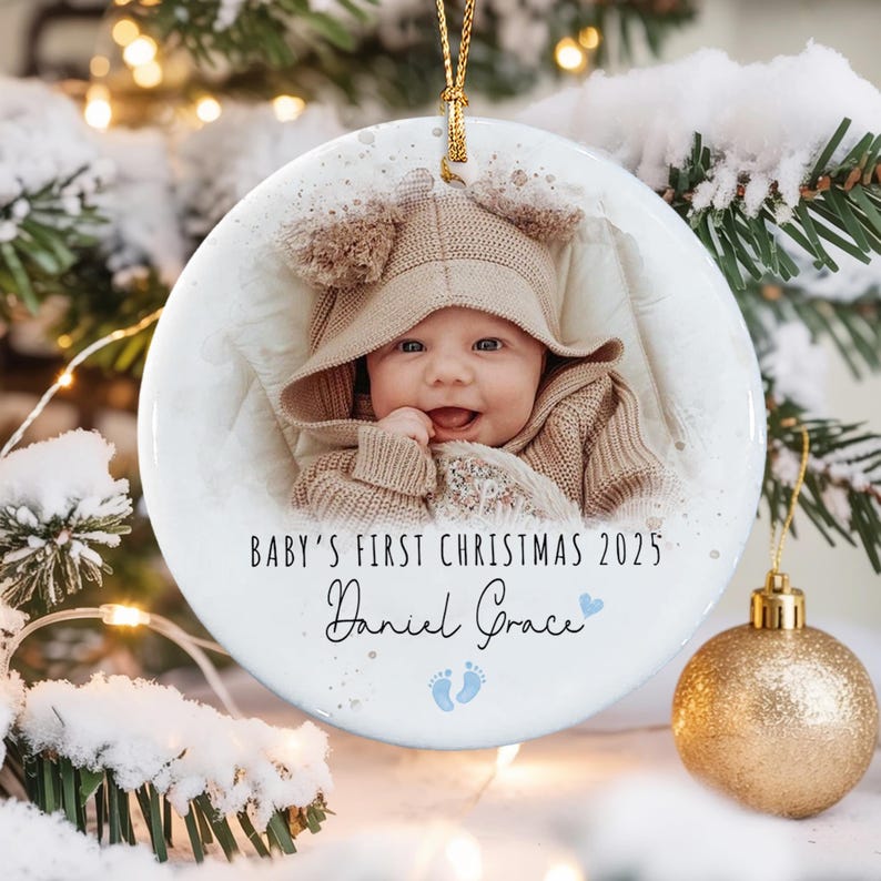 A round white ceramic ornament featuring a photograph of a baby wearing a tan hooded sweater. The text reads BABY'S FIRST CHRISTMAS 2025 and Daniel Grace, accompanied by a small blue heart and two blue baby footprint icons.