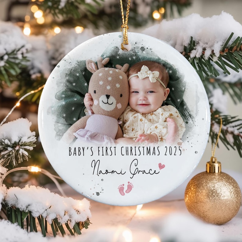 A round white ceramic Christmas ornament featuring a photo of a baby with a plush deer toy. The text reads Baby's First Christmas 2025, the name Naomi Grace, and a small pink heart and baby footprint graphic.