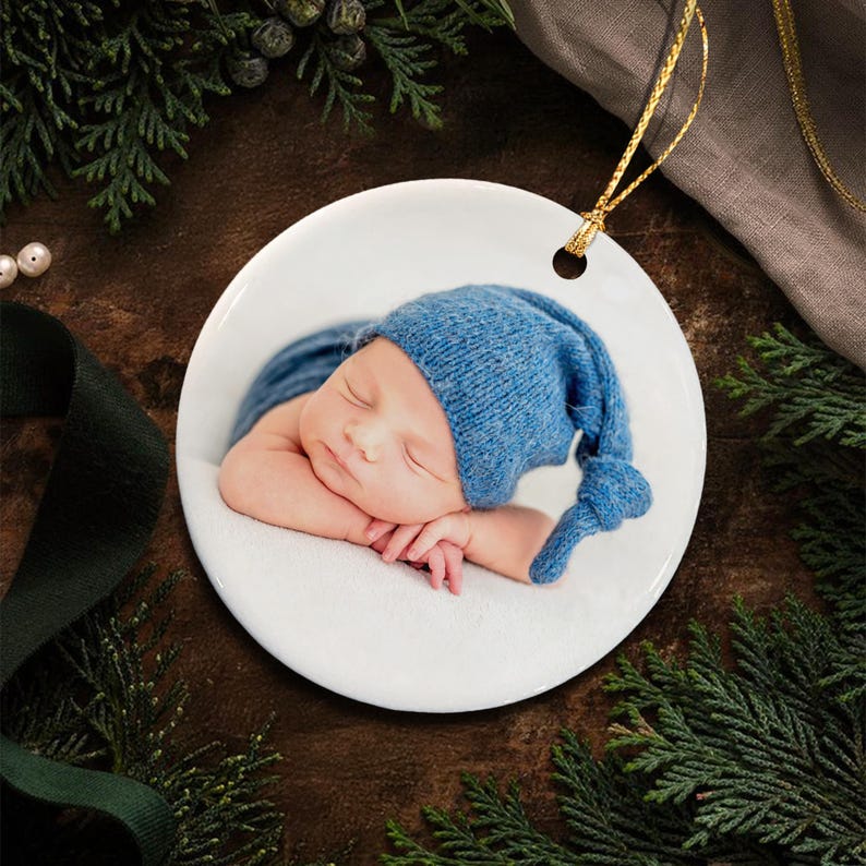 A round white ceramic ornament featuring a printed photograph of a sleeping infant wearing a blue knit cap with a knotted tail.