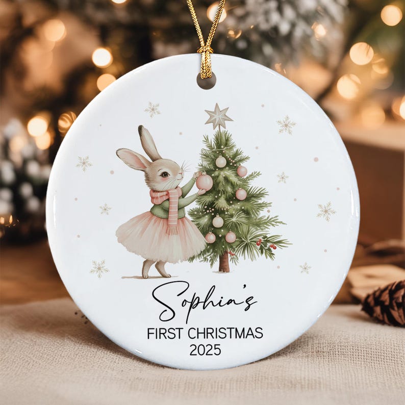 A round white ceramic Christmas ornament featuring a rabbit wearing a pink tutu and scarf, decorating a green Christmas tree with pink ornaments.