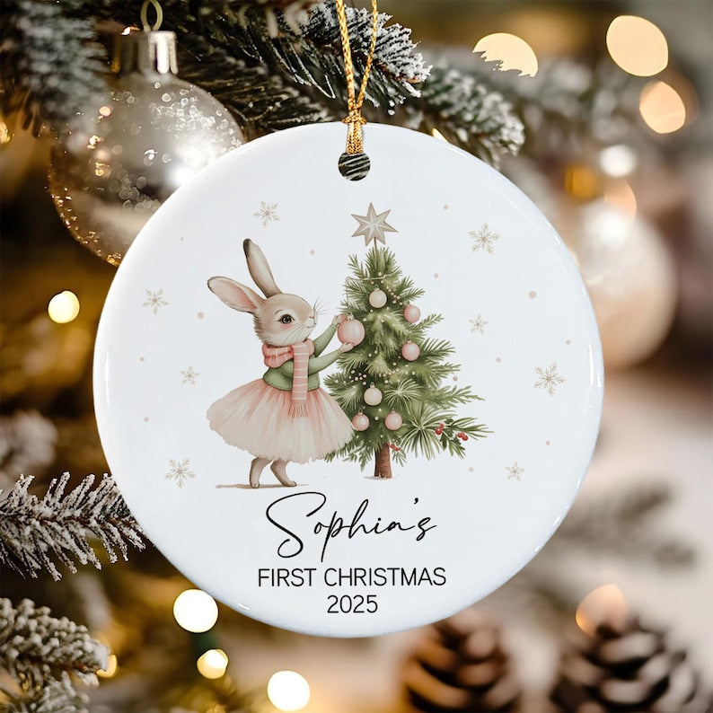 A round white ceramic Christmas ornament featuring a cartoon rabbit wearing a pink scarf and tutu, decorating a green pine tree with pink ornaments. The text reads Sophia's FIRST CHRISTMAS 2025 in black script and block lettering.