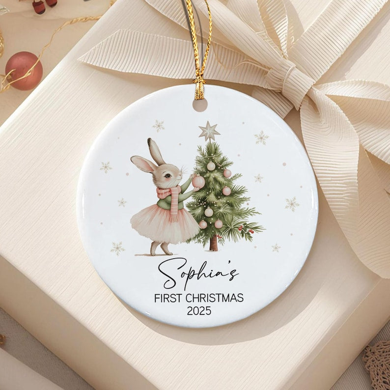 A round white ceramic ornament featuring a rabbit wearing a pink scarf and tutu, decorating a green Christmas tree with pink ornaments. The text reads Sophia's FIRST CHRISTMAS 2025.