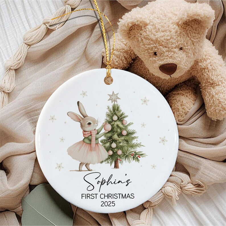 A round white ceramic ornament featuring a rabbit wearing a pink skirt and green sweater decorating a Christmas tree. The text reads Sophia's FIRST CHRISTMAS 2025. A gold string is attached to the top for hanging the ornament.