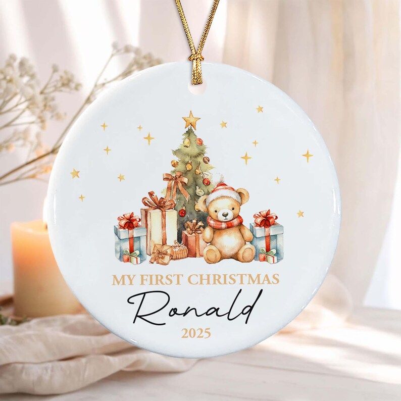 A round white ceramic ornament with a gold string, featuring a watercolor illustration of a teddy bear wearing a red and white hat, a decorated Christmas tree, and gift boxes. Text reads: MY FIRST CHRISTMAS Ronald 2025.