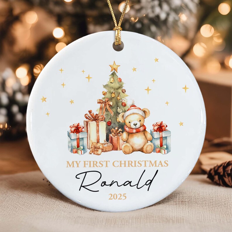 A round white ceramic Christmas ornament featuring a watercolor illustration of a teddy bear wearing a red and white hat and scarf, a decorated Christmas tree, and gift boxes. The text reads MY FIRST CHRISTMAS Ronald 2025.