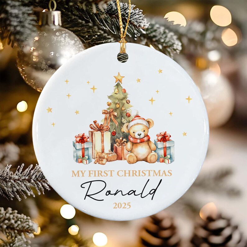 A round white ceramic Christmas ornament featuring a watercolor illustration of a teddy bear wearing a red scarf, a decorated Christmas tree, and gift boxes. The text reads MY FIRST CHRISTMAS, the name Ronald, and the year 2025.