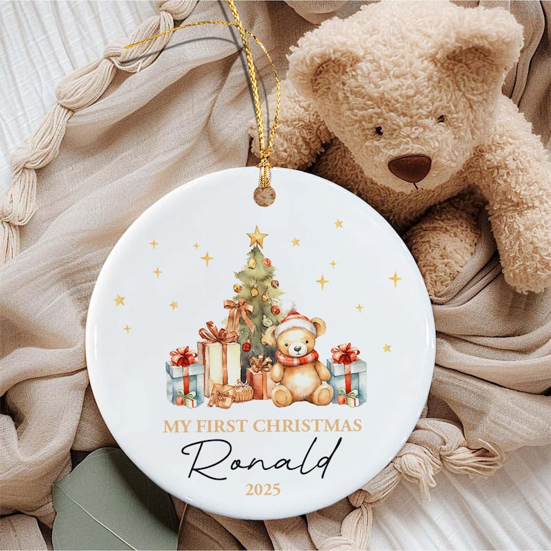 A round white ceramic ornament featuring a watercolor illustration of a teddy bear wearing a red scarf and hat, a decorated Christmas tree, and gift boxes. The text reads MY FIRST CHRISTMAS, the name Ronald, and the year 2025.