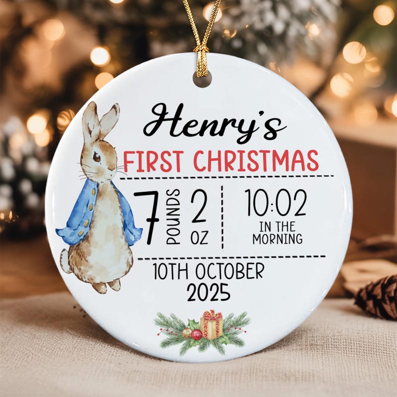 A round white ceramic ornament featuring a watercolor illustration of a rabbit in a blue jacket. Text reads: Henry's FIRST CHRISTMAS, 7 POUNDS 2 OZ, 10:02 IN THE MORNING, 10TH OCTOBER 2025.